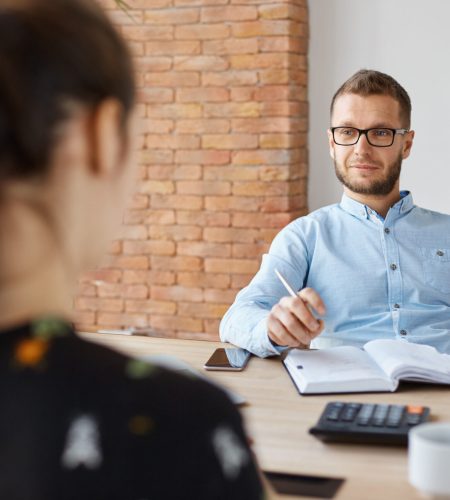 Business concept. Job interview. Adult bearded male hr manager in glasses and shirt sitting in light office in front of brunette caucasian woman, asking questions about previous work place Business concept. Job interview. Adult bearded male hr manager in glasses and shirt sitting in light office in front of brunette caucasian woman, asking questions about previous work place.