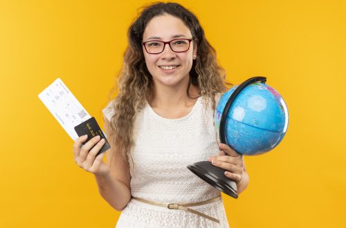 smiling young pretty schoolgirl wearing glasses holding airplane tickets, credit card and globe isolated on yellow background smiling young pretty schoolgirl wearing glasses holding airplane tickets, credit card and globe isolated on yellow background